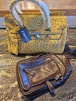 Photo showing two handbags on a table: a yellow ostrich pattern handbag with wrapped handles and lock, a brown leather crossbody bag with front zipper pocket.