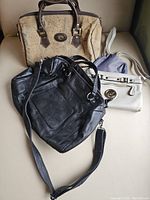 Photo shows four handbags and purses on a beige couch, including a brown fabric handbag with leather trim, a large black leather shoulder bag, a white Michael Kors crossbody purse, and a beige and blue fabric shoulder bag.