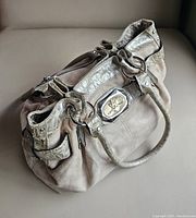 Handbag sitting on a neutral surface showing overall shape, metallic trim, double handles, and front logo plaque.