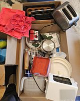 Full overview of cardboard box containing toasters, silicone molds, baking trays, metal strainer, white plates, and kitchen utensils.