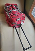 Red rolling duffel bag on a beige couch next to a window, showing telescoping handle pulled out and multiple zipped compartments.