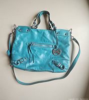 Front view of teal leather Michael Kors handbag with handles, shoulder strap, front zip pocket, and MK logo charm.