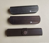 Three eyeglass cases laid out flat, two hard shell Design Optics cases with a dark wood grain design and one separate black perforated soft case with a button flap.
