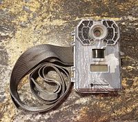 Front view showing the trail camera lens, sensor area, and attached mounting strap on a granite surface