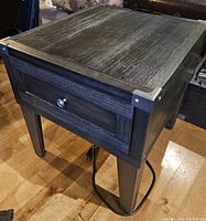 Single dark wood end table with textured wood grain top, single drawer with metal knob, and visible power cable underneath.