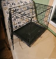 Black wire pet carrier cage with a mesh design, front door open, sitting on a bed with floral quilt.
