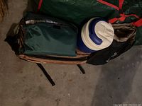 View of camping gear items including green insulated cooler jug, brown Lowepro bag, folded green camping mat, and plaid blanket inside larger carry bag.