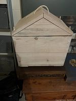 Front and side view of a light-colored wooden chest with a triangular lid and rope handle on top, showing natural wood grain and latch at front.