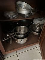 Cookware collection including saucepans, skillets, baking pans, and steamer basket in cabinet shelf