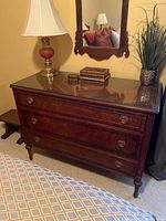 Full view of antique wooden dresser with mirror, three drawers, brass hardware, and items on top including a lamp and decorative boxes.