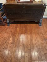 Front view of the antique wooden chest showing full body with carved panels and unique leg design on hardwood floor.
