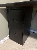 Two-drawer black metal file cabinet side view showing drawers closed under a desk.