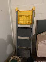 Front view of folding ladder with yellow top platform and three black/gray steps leaning against a wall.