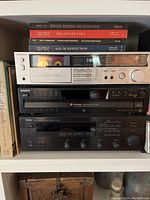 Front view of stacked music system showing Technics cassette deck on top, Sony CD changer in the middle, and Yamaha stereo receiver on bottom with books around.