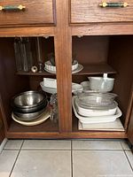 Kitchen cabinet shelf with various stacked pots, pans, bowls, and kitchenware made of metal, ceramic, and glass.