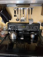 Photo of stove with accessories hanging on rack - ladle, whisk, spatulas, oven gloves, kettle, creamer, and spoon rest visible on stovetop.