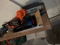 Battery operated chainsaw with an orange handle and 6-inch chain blade, lying on a shelf with charging cables visible.