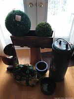 Photo showing the wooden planter box with 2 green plastic topiary balls placed inside it, a wooden rooster decorative statue nearby, 2 blue ceramic planters and a clay pedestal placed on the floor, and a tall metal container with floral designs.
