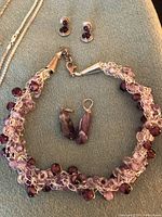 Close up of a silver-tone knotted wire necklace with various amethyst-tone beads and matching pierced earrings with purple stones, also a pair of amethyst drop earrings visible.