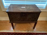 Full view of the wood chest on its stand in front of window blinds.