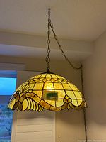 Full view of stained glass swag lamp hanging from ceiling chain, showing domed lampshade in brown and yellow tones and scalloped edges.