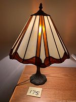 Full lit view of the table lamp showing off-white and brown stained glass shade and ornate metal base on wood surface.