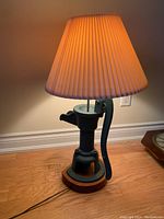 Photo showing full view of cast iron farm pump lamp with pleated shade illuminated, placed on hardwood floor near wall outlet.
