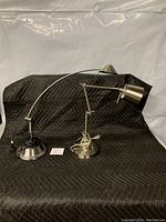 Two chrome finish desk lamps on black quilted surface: one 16-inch LED lamp with curved arm and one 19-inch halogen lamp with articulated arm, both with heavy round bases.