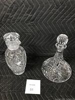 Two cut crystal decanters with pinwheel etching shown side by side on a black quilted surface; one with a wide base and the other taller with pour spouts and stoppers.