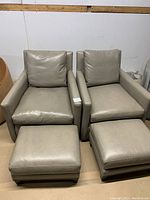 Pair of grey leather swivel chairs with matching footstools placed side by side against a plain background.