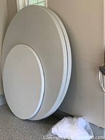Photo shows two large round folding tables leaning flat against a wall, one 6-foot diameter and one smaller, not part of this lot. The 6-foot table is light gray with a white edge trim and folds flat.