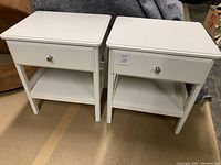 Front view of two white nightstands side by side showing drawer and lower shelf