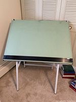 Front view of Alvin drafting table with green vinyl board and black edge lip resting on white metal foldable frame.