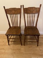 Front view of two antique oak kitchen chairs side by side showing spindled backrests with ornate carved Pauline Johnson motif, turned legs and stretchers, wood grain visible on seats.