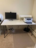 Full view of white office desk with aluminum legs, monitor, printer on extra shelf, set against a beige wall.