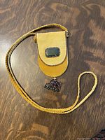 Full view of the yellow hand-made leather pouch with a green jade inset on the flap, detailed beadwork around the flap and pouch base, hanging beaded pendant, and braided leather strap lying on a wooden surface.