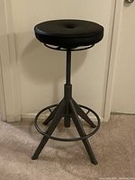 Front view of black adjustable height stool with padded round seat and four metal legs with footrest ring