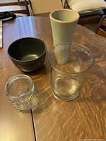 Four vases and bowls arranged on a wooden table, showing light green tall ceramic vase, dark green bowl, clear glass vase, and small textured glass jar.