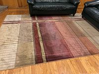 Full top view of DaVinci area rug on hardwood floor between black leather couches showing pattern and colors.