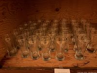 Photo showing rows of 34 clear tall beer glasses arranged on a wooden surface, highlighting their uniform size and shape.