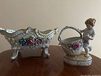 Front side view of both the footed bowl and cherub figurine basket showing floral details and craftsmanship.