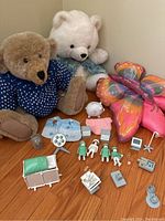 Overall assortment of stuffed animals and Playmobil medical playset items on a wood floor, showing 2 teddy bears, butterfly pillow, 4 figures, medical equipment, and the piggy bank symbolically in the background.