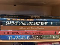 Seven hardcover books stacked horizontally, titles including Renoir, Hollywood Hunks & Heroes, Land of the Inland Seas, A History of the Diocese of Hamilton, The Divine Comedy, and The World's Best Golf.