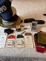 Full view of men's accessories featuring hats, pocket squares in wraps, money clip, cufflinks in boxes, small knife, handkerchiefs on white fabric background.