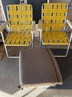 Two vintage folding outdoor chairs side by side with yellow woven webbing seats and backs, aluminum frames, and wooden armrests next to a black mat.
