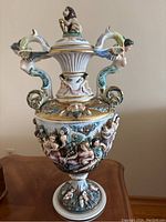 Full view of the tall Capodimonte vase with lid, showing ornate sculpted handles with human figures, lion topper and detailed body decoration.