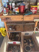 Wooden workbench with assorted tools, tins, adhesives, and hardware items on top, with boxes containing tools underneath.