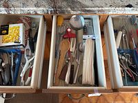 Three open kitchen drawers filled with assorted utensils including knives, spoons, peelers, graters, whisks, ladles, and other cooking tools.