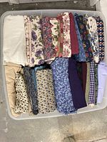 Plastic bin filled with a variety of neatly folded fabric remnants. Fabrics show various floral, solid, and patterned designs in multiple colors.