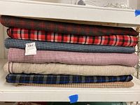 Stack of nine bolts of wool fabric in various plaids, houndstooth, and solid weave patterns arranged on a shelf.